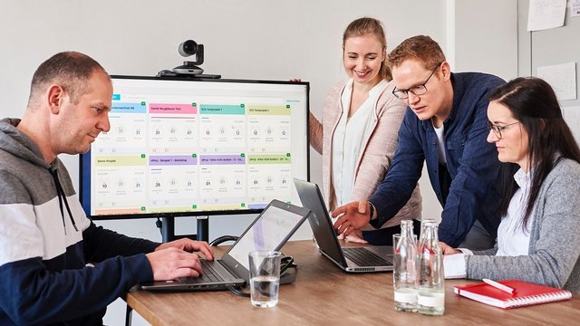 Mehrere Menschen sitzen in einem Konferenzraum und arbeiten. Ein Mann zeigt einer Frau etwas am Laptop.