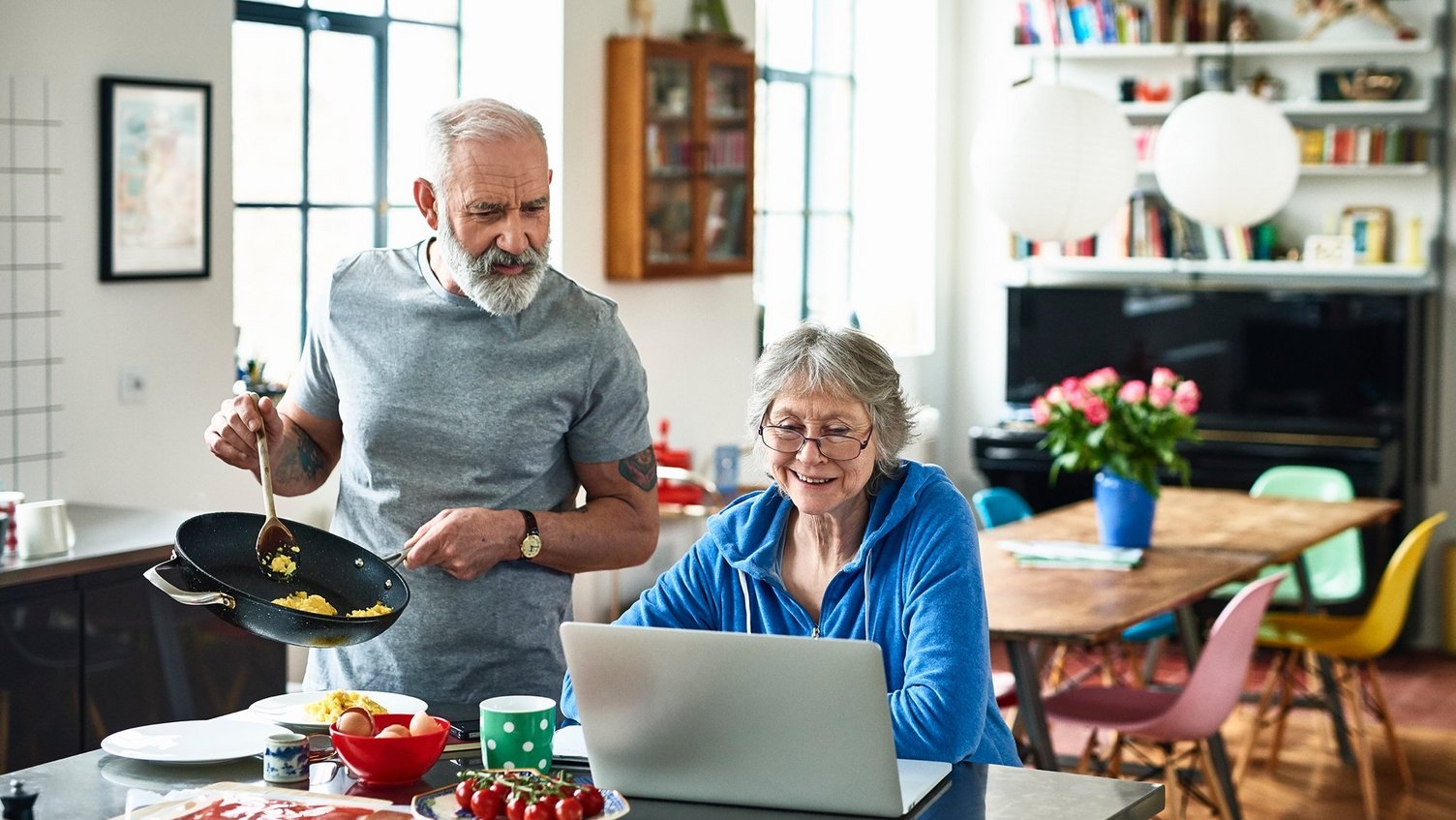 Ein älteres Ehepaar in der Küche. Die Frau sitzt lächelnd an ihrem Laptop. Der Mann bereitet das Essen zu und schaut ihr interessiert über die Schulter.