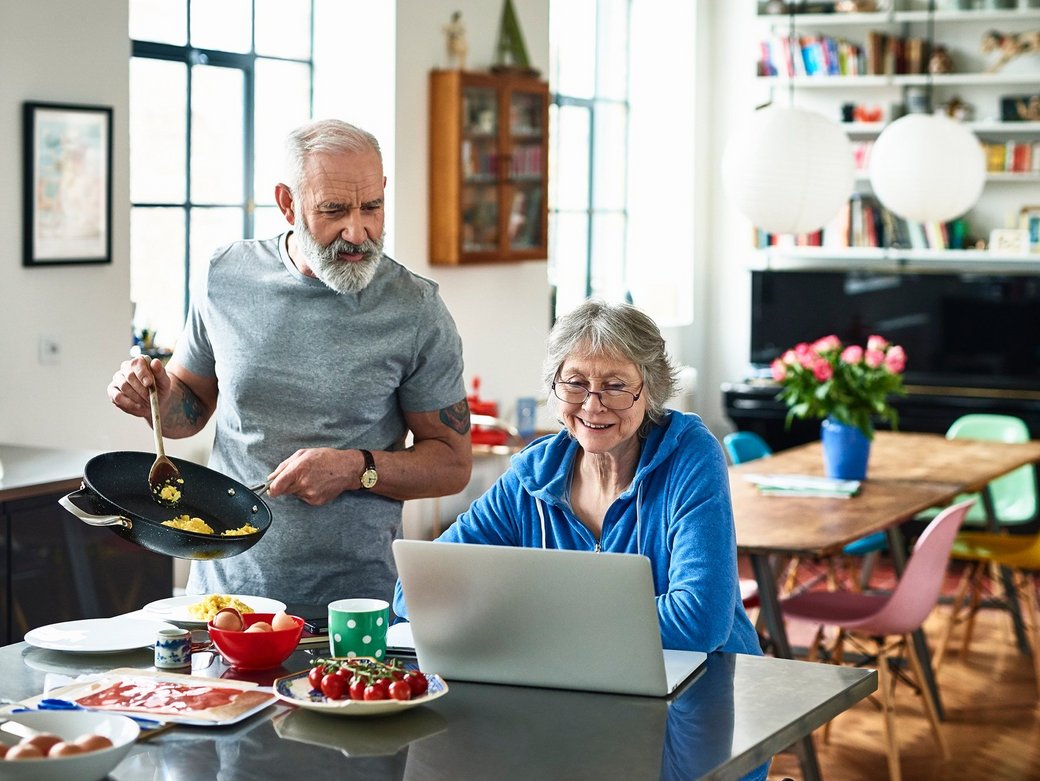 Ein älteres Ehepaar in der Küche. Die Frau sitzt lächelnd an ihrem Laptop. Der Mann bereitet das Essen zu und schaut ihr interessiert über die Schulter.