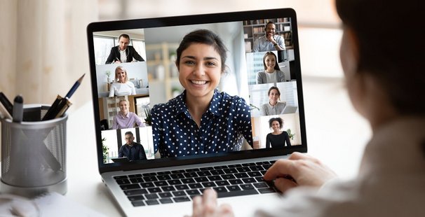 Eine Frau sitzt am Schreibtisch und schaut auf einen Computer-Bildschirm auf dem mehrere Teilnehmer*innen per webcam teilnehmenIndian ethnicity young woman lead video call distant chat, group of different mates using videoconference