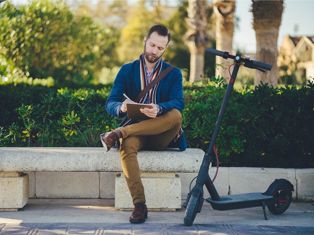 Ein Mann mit E-Roller sitzt draußen auf einer Bank und notiert etwas auf seinem Tablet