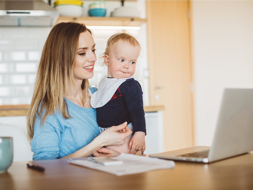 Eine junge Mutter sitzt an ihrem Laptop mit hält ein Baby im Arm.