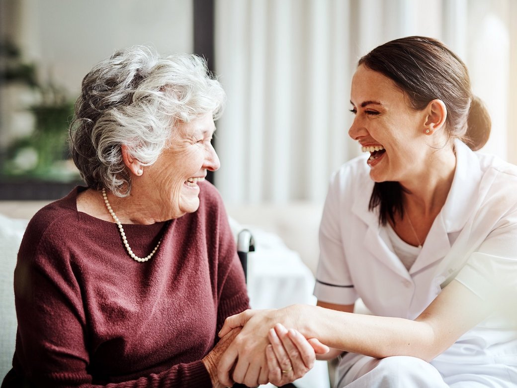Ältere Dame hat Besuch von einer Pflegekraft. Sie halten sich an der Hand und lachen gemeinsam herzlich.