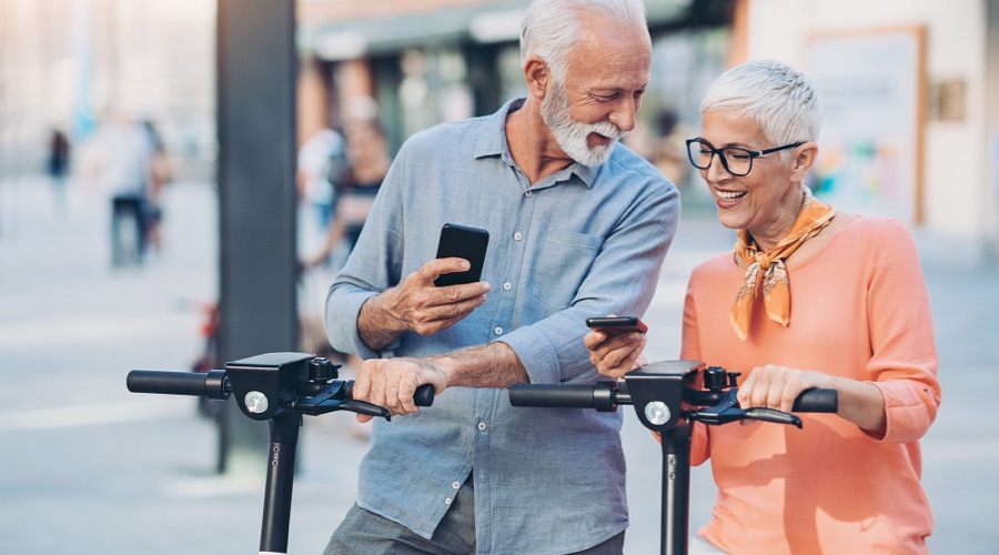 Zwei moderne Senioren auf Elektro-Rollern. Sie lachen und schauen interessiert auf ihre digitalen Endgeräte.